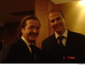 This is a photograph showing two men in dark suits and ties at what appears to be an indoor social or formal event. The image was taken on a date stamp showing '2' and time '7:18AM', suggesting early morning hours. The photo quality and color tone in