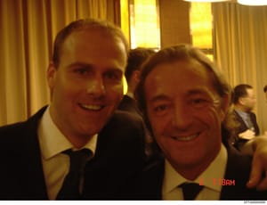 A candid photograph taken at what appears to be a formal indoor event or gathering, showing two men in business suits smiling at the camera. The photo is dated 2-7-18 (February 7, 2018) based on the timestamp visible in the lower right corner. The se