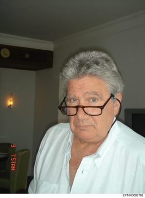 This is a portrait photograph of an older male subject with gray hair and glasses, taken indoors in what appears to be a residential setting. The photo bears an evidence identifier stamp and Bates number consistent with investigative documentation. T