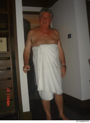 This is an evidentiary photograph showing a middle-aged male subject in an indoor residential hallway or corridor, wrapped in a white towel. The image appears to be taken as part of investigative documentation, with a visible date stamp and evidence 