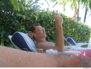 This is a candid photograph showing a man relaxing at what appears to be a poolside or beach setting with tropical vegetation in the background. The image was taken on June 20th at 2:57 PM (based on the timestamp visible). The photograph appears to b