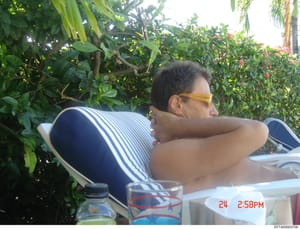 This is a casual photograph showing a person seated outdoors in a garden or landscaped setting with tropical vegetation visible in the background. The image appears to be from a social gathering or meal, with beverage glasses and dishware visible in 
