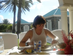 This is a casual photograph taken at an upscale residential property showing a woman seated at an outdoor dining table. The image captures the luxurious setting of what appears to be a private estate with palm trees, manicured grounds, and classical 