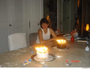 This is a casual photograph of a birthday celebration showing a person seated at a dining table with lit birthday cakes in front of them. The image appears to have been taken in an upscale residential interior space, with white cabinetry visible in t