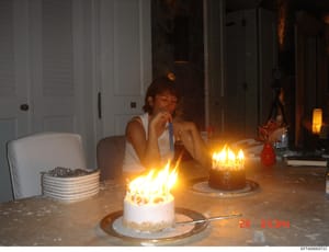 This is a casual photograph showing an interior domestic scene with a person seated at a table with lit birthday cakes. The image appears to be from a personal collection or surveillance file, dated in the early 2000s based on the timestamp visible (