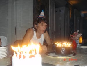 This is a personal photograph showing a young woman at a birthday celebration, wearing a blue 'Happy Birthday' party hat and white tank top, blowing out candles on birthday cakes. The image appears to be from a domestic or residential setting and is 