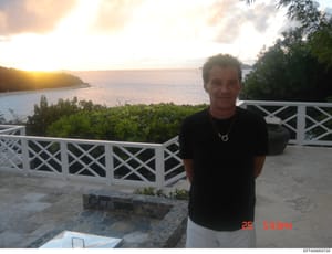 A personal photograph showing a middle-aged man standing on a beachfront deck or patio area with ocean and tropical vegetation in the background. The image appears to be from a residential property in a tropical location, likely taken sometime in the