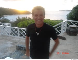 A casual photograph showing a person standing on a deck overlooking a water view with tropical vegetation. The image is timestamped 25 5:49PM and appears to be taken during sunset or golden hour. The setting suggests a residential property with signi