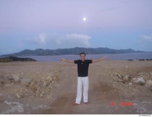 This is a casual photograph showing a man posing on a rocky beach with his arms outstretched. The image captures a waterfront location with distinctive mountains and a developed hillside cityscape visible across the water. The photo is timestamped Ju