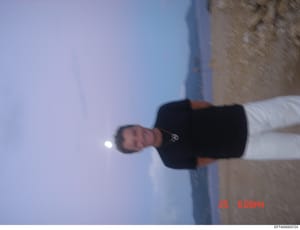 This is a candid photograph showing a person standing outdoors against a clear blue sky and stone/concrete wall background. The image is rotated 90 degrees and dated 25 June at 6:06 PM based on the timestamp visible in the lower right corner. The pho