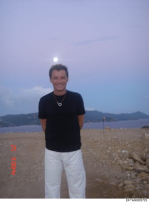 This is a casual personal photograph showing a man standing on a beach at dusk or dawn with a full moon visible in the sky. The image appears to be from the early 2000s based on photo quality and style. The location features a sandy beach with rocky 