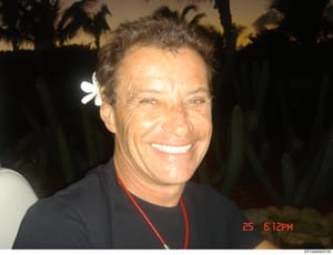 This is a personal photograph showing a middle-aged man smiling at the camera during an evening event in a tropical setting. The photo is dated 25 (month unknown) at 6:12 PM and includes palm trees silhouetted against the night sky in the background.