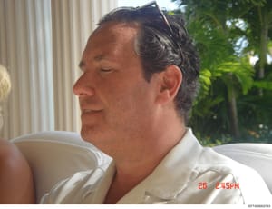 This is a casual photograph of a middle-aged male subject taken outdoors, dated 26 (month unknown) at 2:45 PM based on the timestamp visible in the lower right corner. The photo shows the subject in profile wearing a white/cream colored shirt, seated
