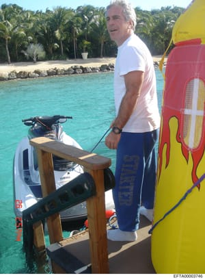This is a photograph showing a middle-aged man in casual attire (white t-shirt, blue shorts) standing on what appears to be a dock or boat platform in tropical waters. The scene includes a Yamaha jet ski, windsurfing equipment, and a palm tree-lined 