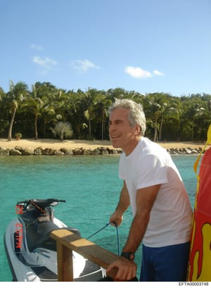 This is a personal photograph showing a middle-aged man with gray hair wearing a white t-shirt and dark shorts, positioned at the helm of a jet ski in clear turquoise water. The setting appears to be a tropical location with palm trees, sandy beach, 