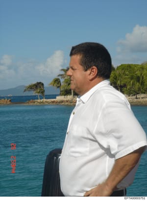 This is a photograph showing a man in a white short-sleeved shirt photographed in profile at what appears to be a tropical coastal location. The image shows a waterfront scene with palm trees, clear blue water, and mountainous terrain in the backgrou