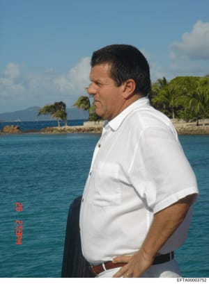 This is a photograph of a middle-aged male wearing a white short-sleeved shirt, standing in profile against a tropical waterfront backdrop. The image appears to be taken during daytime with clear skies and palm trees visible in the background. The ph