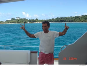 This is a personal photograph showing an unidentified man on the deck of a boat in tropical waters, with a beach and tree-lined coastline visible in the background. The image is timestamped as taken on the 26th at 3:05 PM. The photo appears to be inc