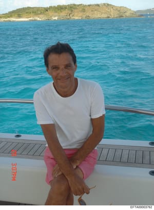This is a personal photograph showing a middle-aged man posing on the deck of a yacht in clear tropical waters. The image appears to be evidence documentation from the investigation, marked with a Bates number and archive stamp. The photo captures th