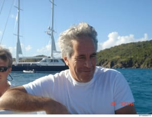 This is a casual photograph showing a man with white/gray hair wearing a white t-shirt on what appears to be a boat or yacht in tropical waters. A large sailing yacht is visible in the background, along with forested coastline. The image is dated 26 