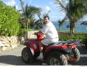 This is a personal photograph showing an unidentified man sitting on a red all-terrain vehicle (ATV) at what appears to be a tropical coastal property. The image was taken on March 10 (based on timestamp) at 3:30 PM and shows the subject in casual at