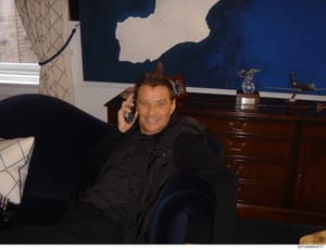 This photograph shows an unidentified man in what appears to be a formal residential interior, speaking on a telephone while seated on a dark-colored sofa. The room features modern artwork with blue and white abstract elements, a wooden dresser with 