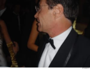 This is a photograph showing a man in profile wearing formal evening attire (tuxedo with bow tie and white dress shirt) at what appears to be a formal event or social gathering. The image is a close-up side view with other people visible in the backg