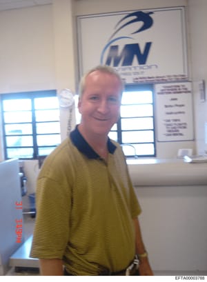This is a candid photograph of a middle-aged male with reddish-blonde hair wearing a yellow striped polo shirt, taken indoors at what appears to be an aviation-related facility or office. A sign bearing an aviation company logo (appearing to show 'MN
