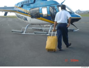 This is a photograph showing a blue and yellow helicopter (registration N484GH visible) parked at an airfield with a person in light blue shirt and dark pants carrying a yellow suitcase approaching the aircraft. The image is dated 31 at 3:50 PM and d