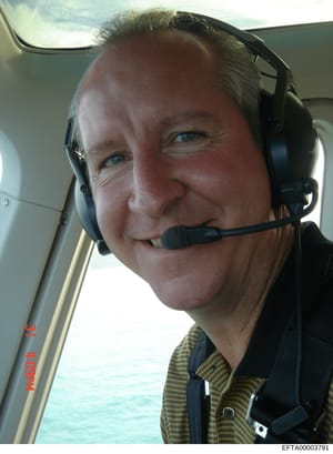 This is a personal photograph taken inside an aircraft cockpit showing a male pilot wearing a communication headset. The image appears to be a selfie taken during flight, with a window visible showing sky and water below. The photograph's inclusion i