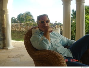 This is a personal photograph showing a man wearing sunglasses and a light blue shirt, seated in a wicker chair on a covered patio or colonnade. The setting features classical white columns, tropical vegetation including palm trees, and stone walls i