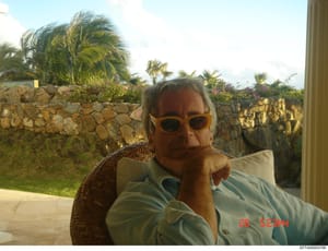 This is a personal photograph showing a middle-aged man wearing orange-tinted sunglasses and a light blue shirt, seated outdoors on what appears to be a wicker chair. The setting is a tropical property with a stone perimeter wall, manicured landscapi