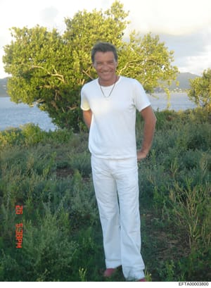 This is a casual personal photograph showing a middle-aged man wearing all-white casual clothing (t-shirt and pants) standing outdoors in what appears to be a Mediterranean or tropical coastal setting. The image includes a large green tree, Mediterra