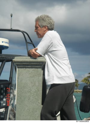 This is a photograph depicting a middle-aged man with gray hair wearing a white shirt and dark pants, standing on or near a boat with visible water and coastline in the background. The image appears to be documentary in nature, with a timestamp visib