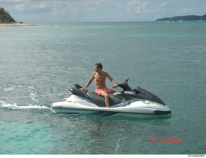 This is a photograph showing a shirtless young male sitting on a white and black jet ski in clear turquoise ocean water. The image appears to be dated 07/12/54pm based on the red timestamp visible in the lower right corner. The tropical setting with 