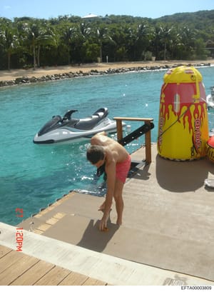This photograph shows a private waterfront property featuring a wooden dock with a jet ski, inflatable water toys, and a person in the water. The property includes a sandy beach with palm trees and dense vegetation on a hillside in the background. Th