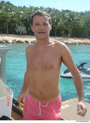This is a personal photograph showing a middle-aged man standing in shallow tropical waters near a dock or pier. The image appears to be from a vacation or leisure setting, with a jet ski visible in the background and palm trees lining a sandy beach.