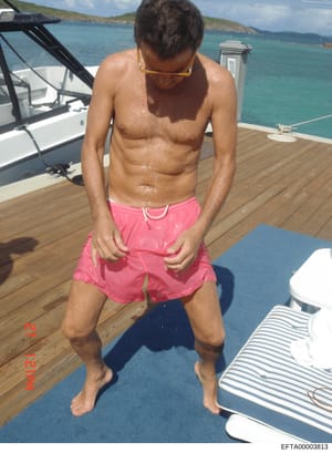 This is a personal photograph taken on the deck of a yacht, showing a shirtless male subject wearing pink swim shorts and yellow-tinted sunglasses. The image depicts a tropical Caribbean setting with turquoise water, green hillsides, and moored vesse