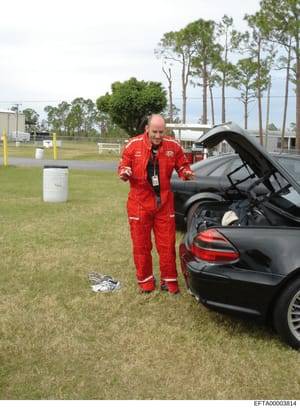 This photograph shows a man in full red firefighter protective gear and an ID badge standing next to a black sedan with its hood open at what appears to be a training or demonstration facility. The image includes industrial storage tanks, grass field