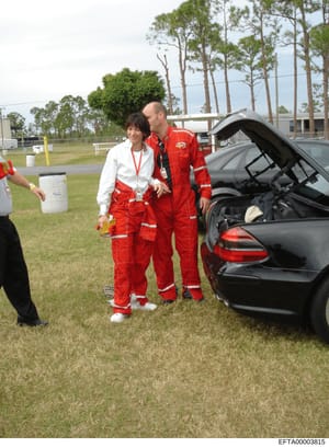 This photograph shows two individuals at what appears to be an emergency response or hazmat training event. A woman in a white jacket and red protective suit stands next to a male instructor or official also wearing a red protective suit with reflect