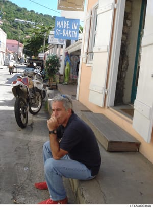 This photograph shows Jeffrey Epstein crouching on a street in Saint Barthélemy (St. Barth), a Caribbean island location. The image captures him in a casual pose wearing a navy polo shirt and jeans on what appears to be a commercial street with local