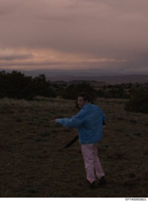 This is a photograph showing a man standing in an arid desert landscape during twilight hours. The man wears a blue jacket and light-colored pants and appears to be gesturing or pointing. The image is part of the Epstein investigation files but lacks