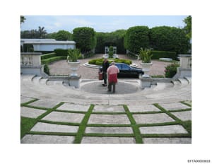 This photograph depicts the front entrance courtyard of a large residential property, featuring a circular driveway with ornamental landscaping, geometric paving patterns with grass inlays, decorative urns, and extensive manicured hedging. Two figure