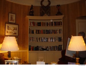 This photograph shows the interior of an upscale residential study or library room featuring floor-to-ceiling bookshelves, decorative African sculptures, framed artwork, and period lighting fixtures. The room displays wealthy home furnishings and art