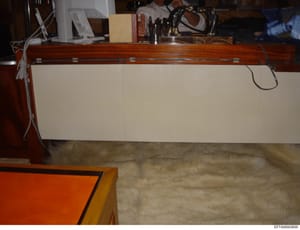 This is a photograph of an interior residential space showing a wooden desk or work surface with a cream-colored drawer front, various office items and personal effects on top, and what appears to be a carpeted floor. The image appears to be document