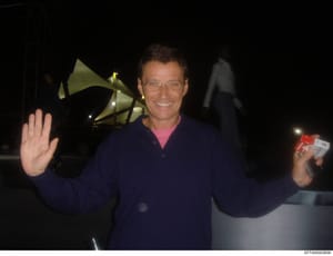 This is a personal photograph taken at night showing a man wearing glasses and dark clothing with a pink undershirt, waving at the camera while holding what appears to be a red object. The setting appears to be an outdoor facility with large satellit