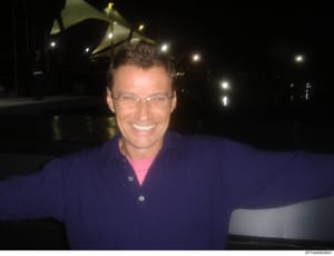 This is a photograph of a man wearing glasses and a dark blue jacket with a pink shirt underneath, photographed at night in front of what appears to be a private aircraft or hangar with illuminated fixtures visible in the background. The image is inc