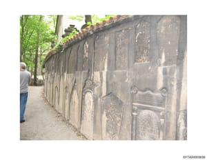 This is a photograph showing a long stone cemetery wall or mausoleum facade with multiple ornamental plaques and burial inscriptions. The image captures weathered stone surfaces with decorative scrollwork and engraved memorial plates. A person in cas