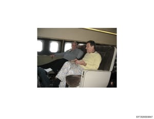 This photograph shows two men seated in the luxury interior of a private aircraft. The image appears to be documentary evidence capturing individuals aboard a private jet, with upholstered seating and cabin windows visible. The photo is potentially r