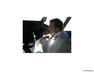 This is a photograph showing a man in the cockpit of an aircraft, appearing to be at the controls or operating equipment. The image shows the interior of what appears to be a private aircraft with visible cockpit instrumentation. The photo is evidenc